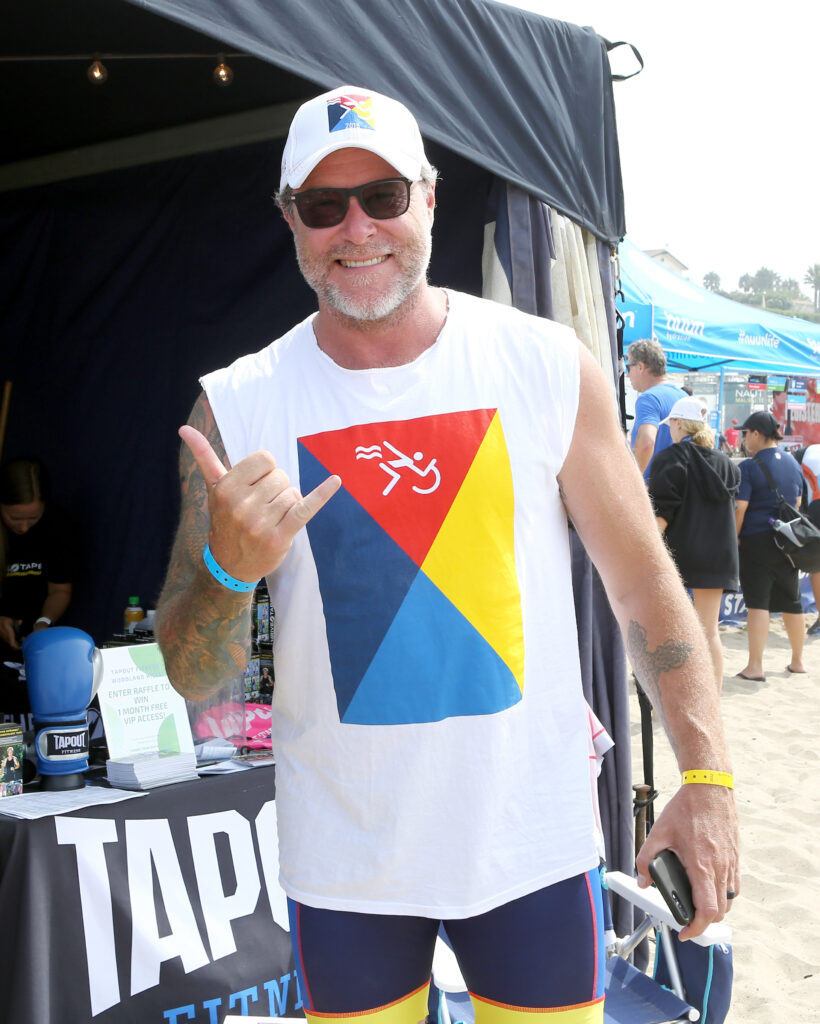 Dean McDermott smiles while wearing a sleeveless white shirt and a white ball cap. He is standing outdoors at the 33rd Annual Nautica Malibu Triathlon.