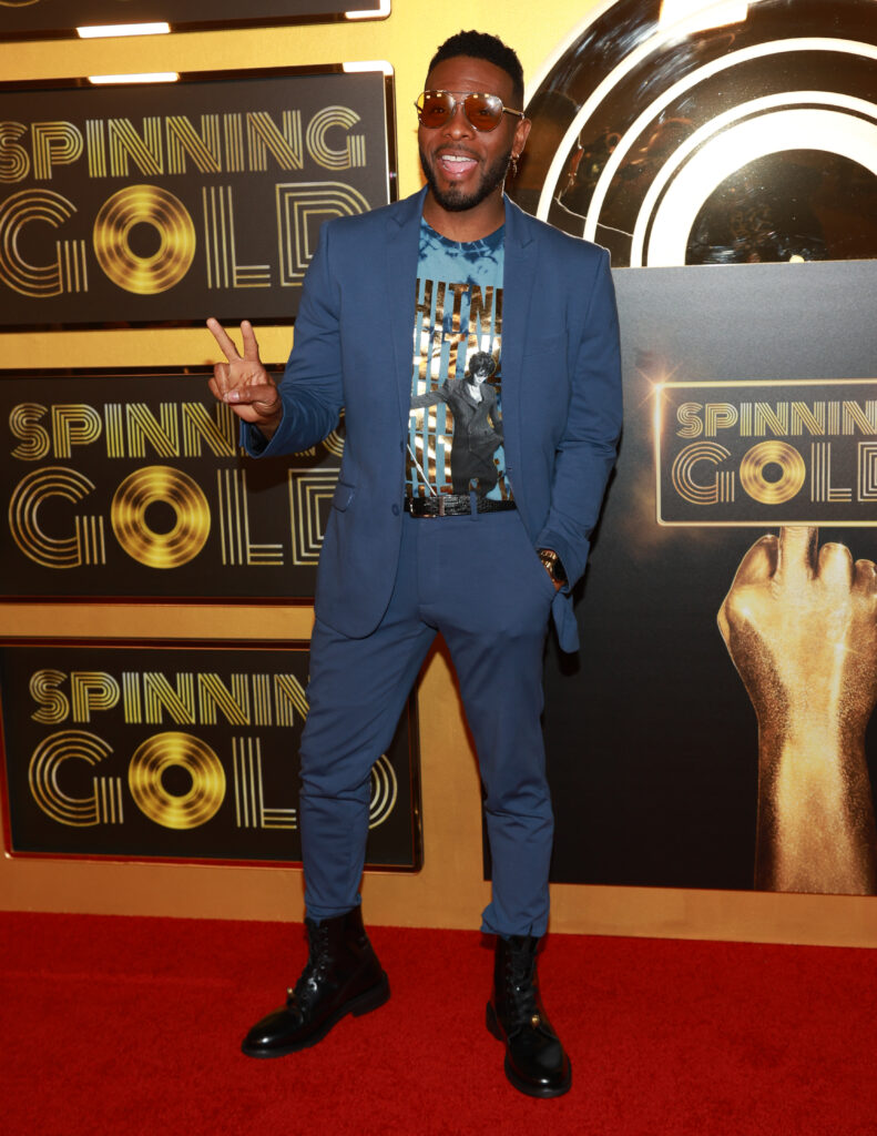 Kel Mitchell stands in front of a gold background at a premiere, wearing a subdued blue suit.