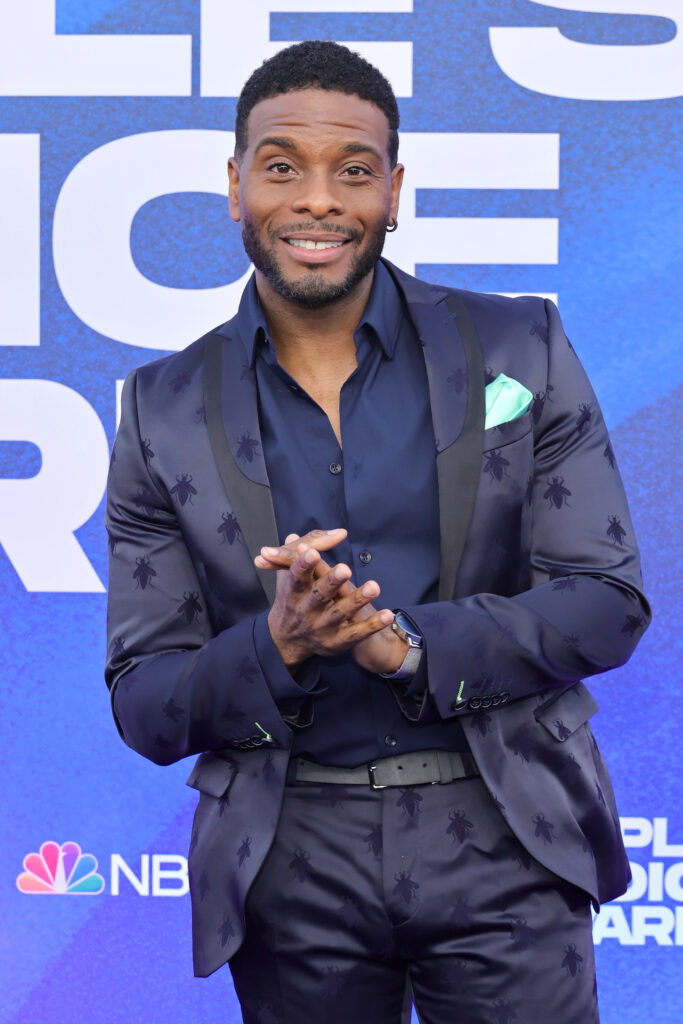 Kel Mitchell poses with his hands clasped expectantly while in a dark and very blue purple ensemble.