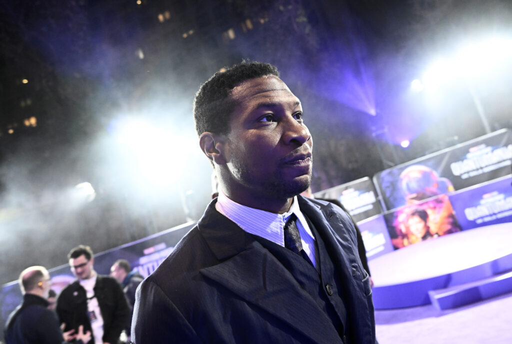 Jonathan Majors walks at a public appearance with colorful posters behind him.