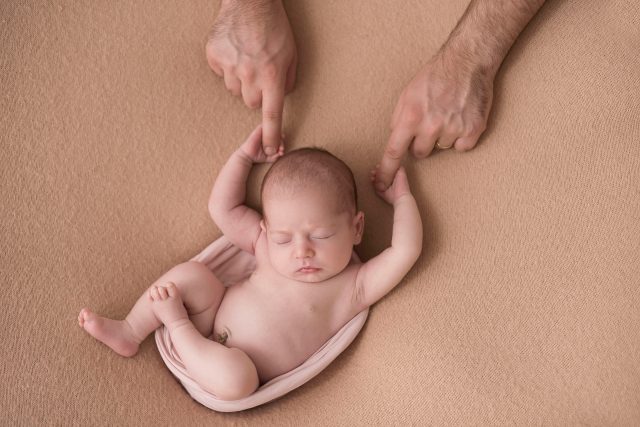 bebê recém-nascida em puff de barriga pra cima com pai segurando as mãos da bebê por cima no estúdio fotográfico laura alzueta