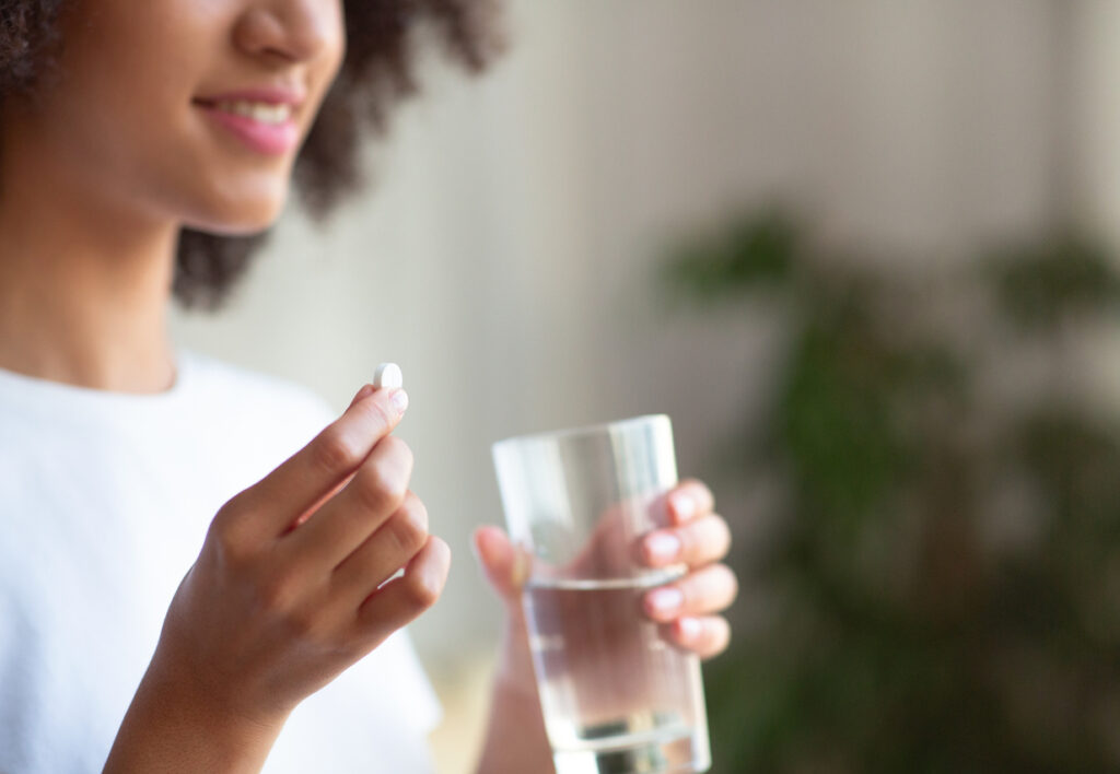 Smiling woman taking white round pill, holding water glass in hand. Happy young female taking supplement, daily vitamins for hair, skin, body, natural beauty and healthy lifestyle, free space, cropped