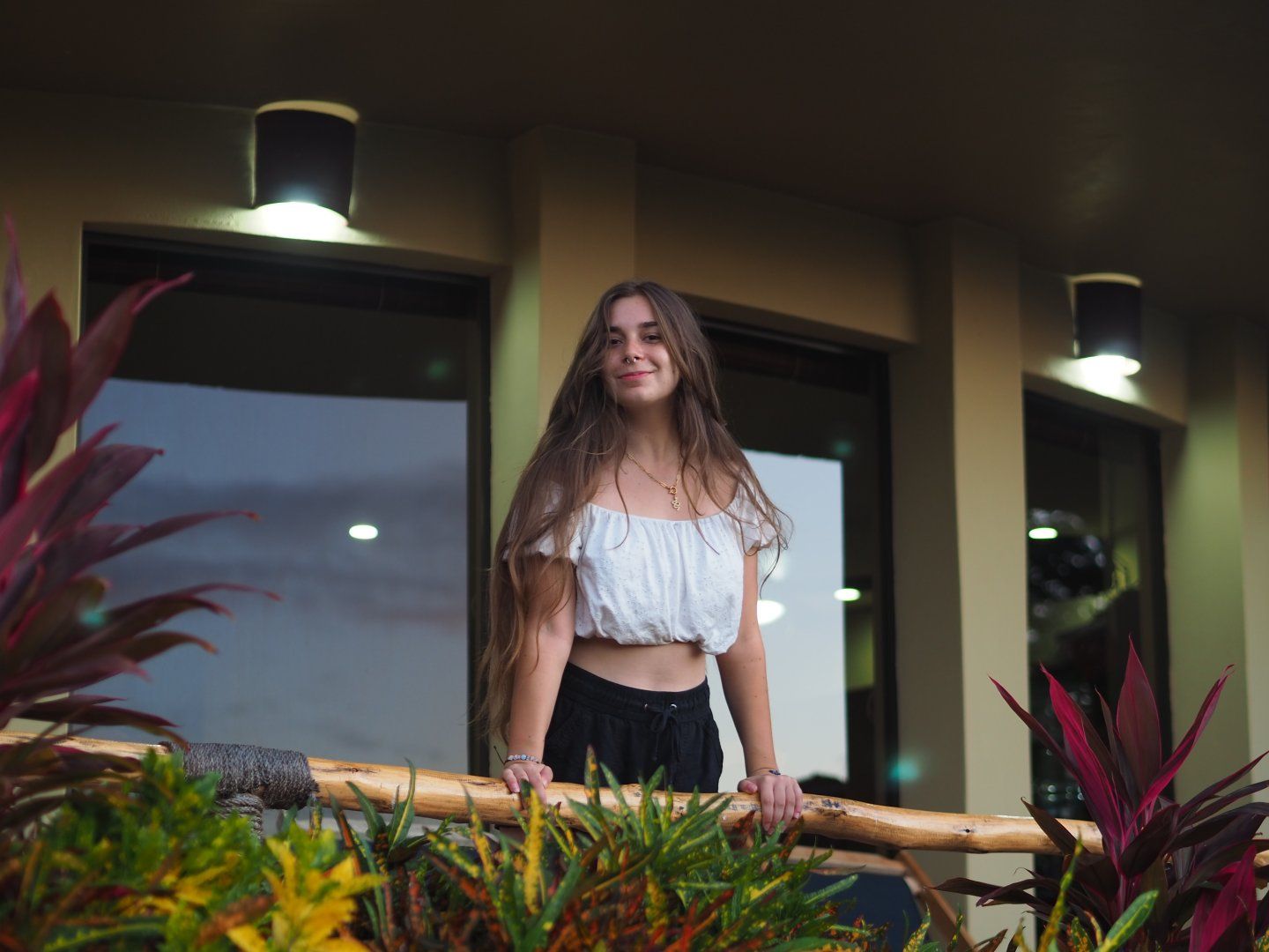 Camille in white top with bare shoulders and bare midriff leaning over balcony overlooking pool at an apartment rental near the beach.