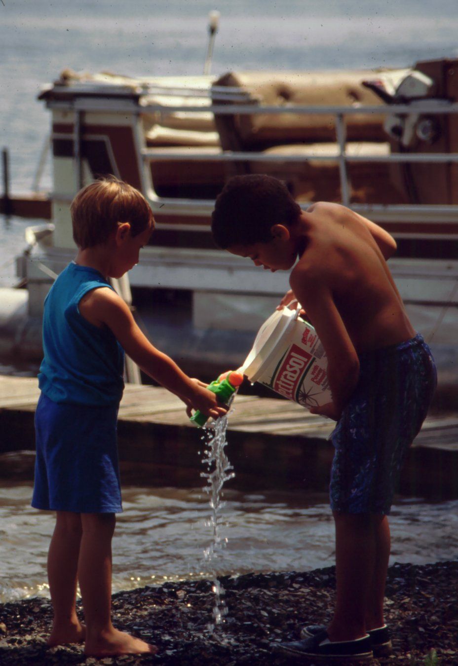 Two kids. One on left holding a squirt fun. One on right pouring water into it from a bucket. Standing on a rocky beach on Conesus Lake in the Finger Lakes region of Western New York summer 1994.