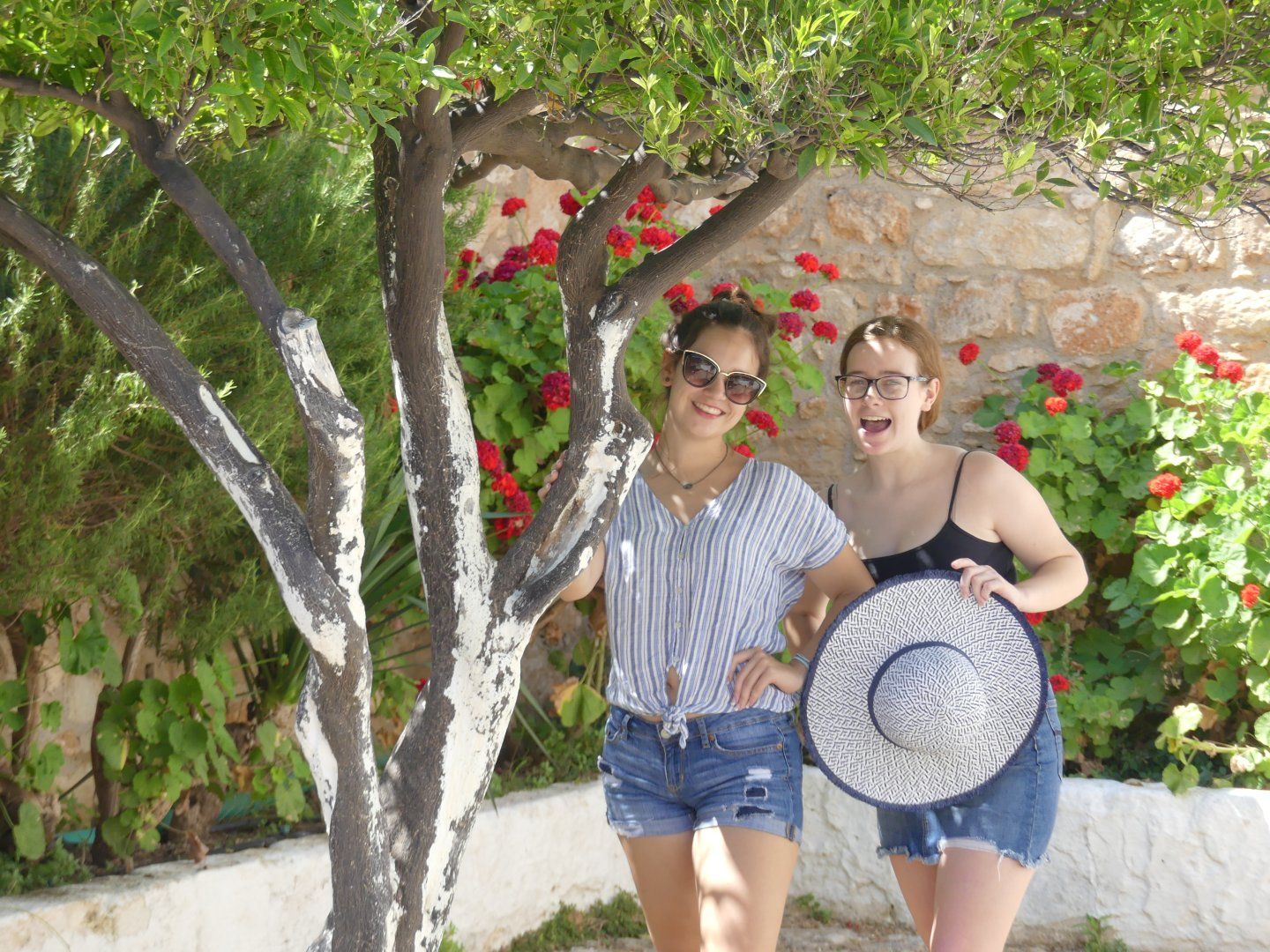 Emily Grice in striped shirt, jean shorts and sunglasses in middle of picture. Manicured tree on left. In the shade of Aptera's courtyard. Madeline on right in black camisole top and jean miniskirt holding a sun hat with glasses.