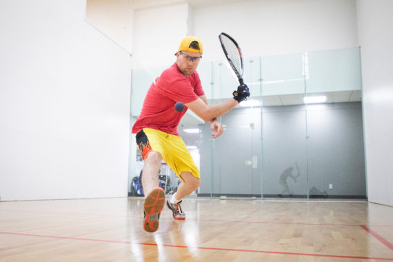 Men's National Team member Lee Connell is hitting a racquetball with his racquet using a backhand swing.