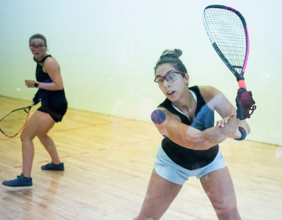 Michèle Morissette is using a back hand swing to strike the raquetball. In the background, her opponent, Juliette Parent is looking over her should towards Morissette as the ball is being hit.