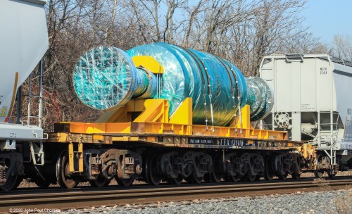 A twelve-axle flat car carries what resembles a gigantic piece of candy but is probably an electrical power plant component, north on the mixed freight, in Jersey City, NJ.