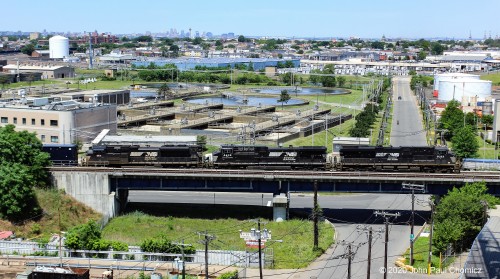 An NS empty trash train heads east towards the Arthur Kill Drawbridge that will take these empties to Fresh Kills landfill, in Staten Island, to be filled. The Newark Skyline and Newark Liberty International Airport control tower are visible in the background.