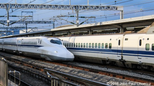 A southbound Shinkansen train makes a station stop in Odawara, Japan as another southbound Shinkansen expresses through the station. Both of these train sets are the N700 series, which is the newest type of train set on that railway.