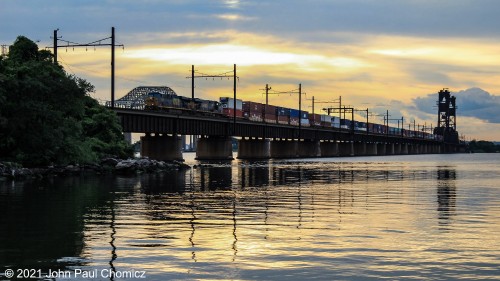 Under a dramatic evening sky, the crossing of an eastbound CSX intermodal train is reflected in the Newark Bay.